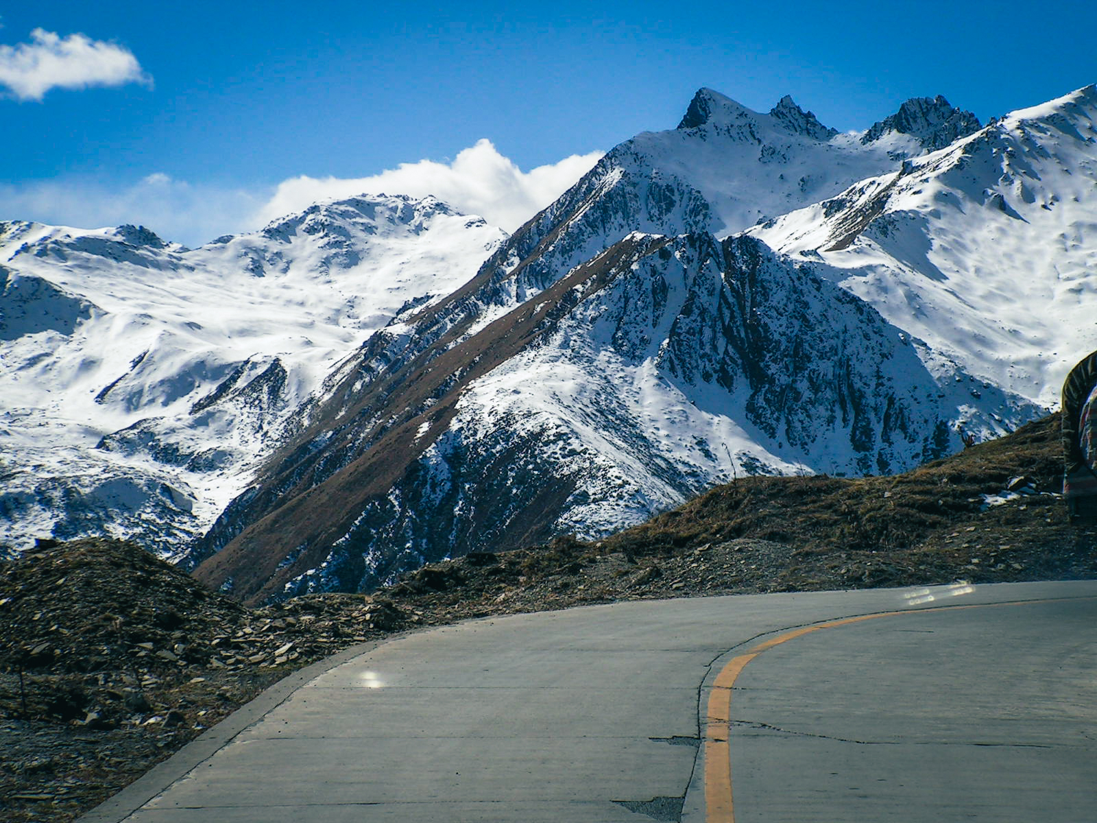 Concrete mountain road curving past snow-covered rocky peaks under a deep blue sky at Balang Pass in the Qionglai Mountains, Sichuan, China, in 2004.