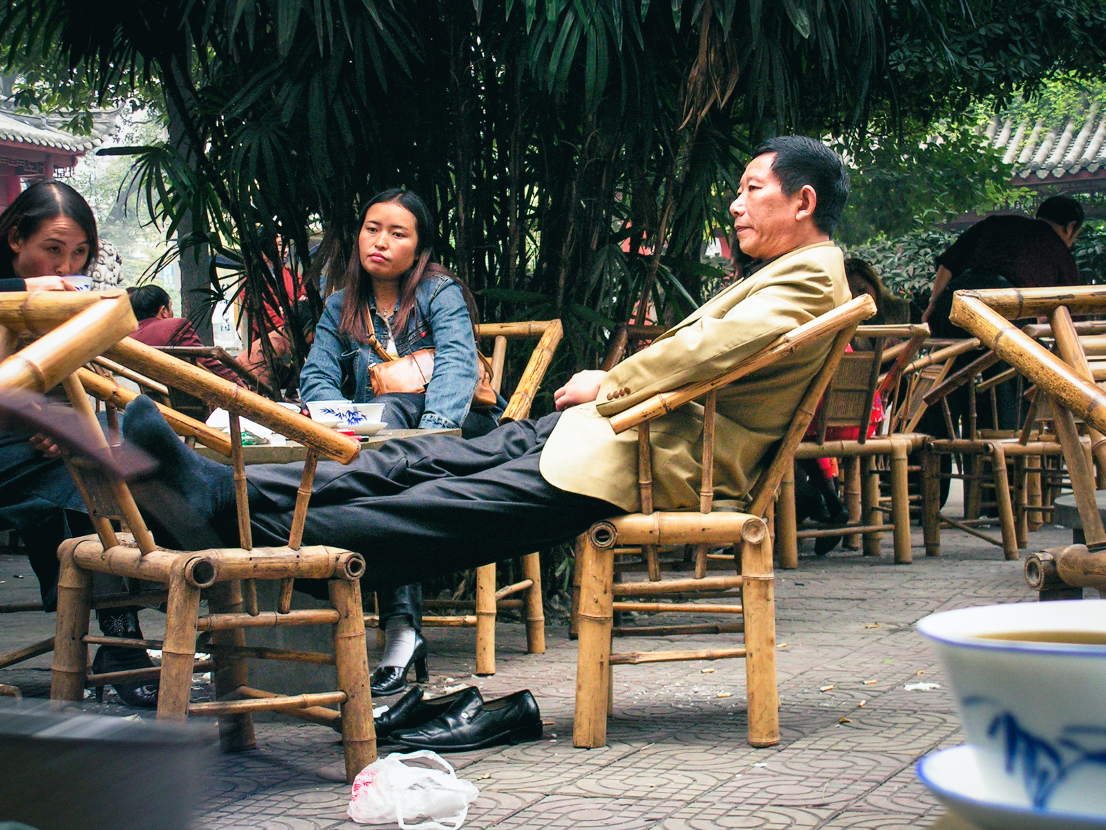 A man in a beige jacket reclining in a bamboo chair with his shoes off and feet up on another chair at an open-air teahouse in Chengdu, China, in 2004, with other guests drinking tea under the trees.