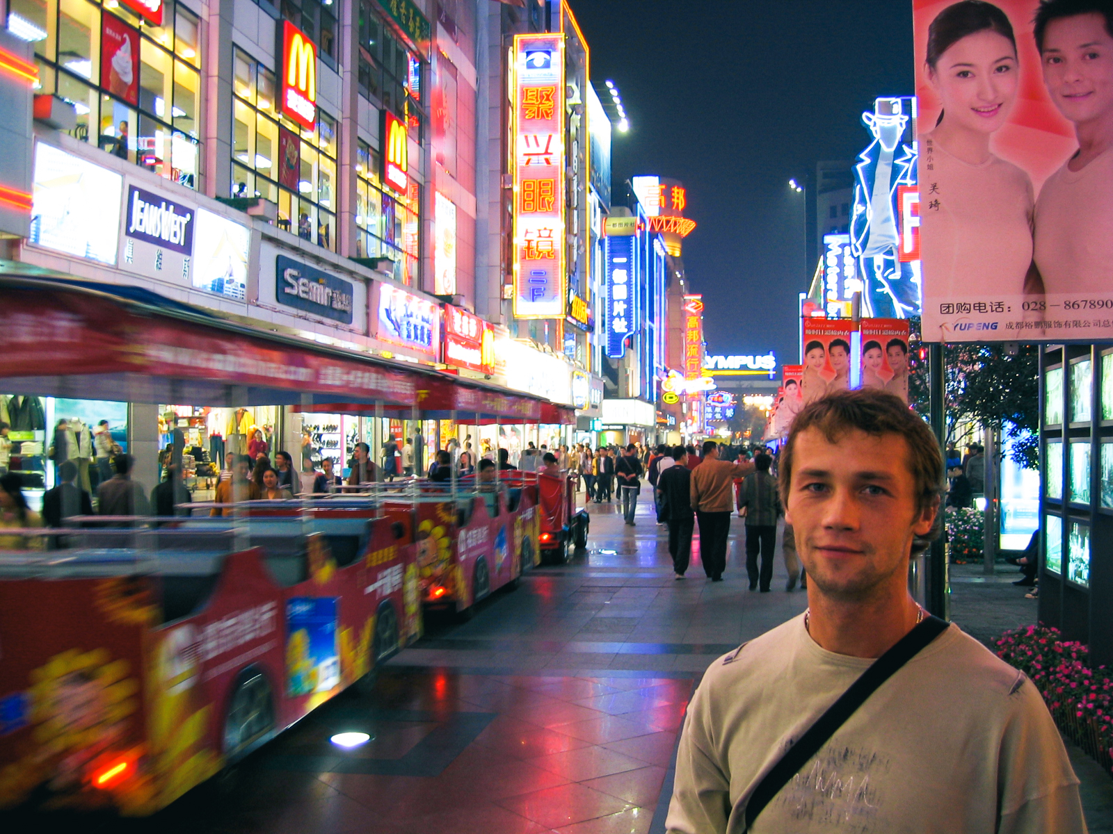 Oleg Gritskevich standing on a neon-lit pedestrian shopping street in Chengdu, China, at night in 2004, with a red sightseeing road train, shop signs and crowds behind him.