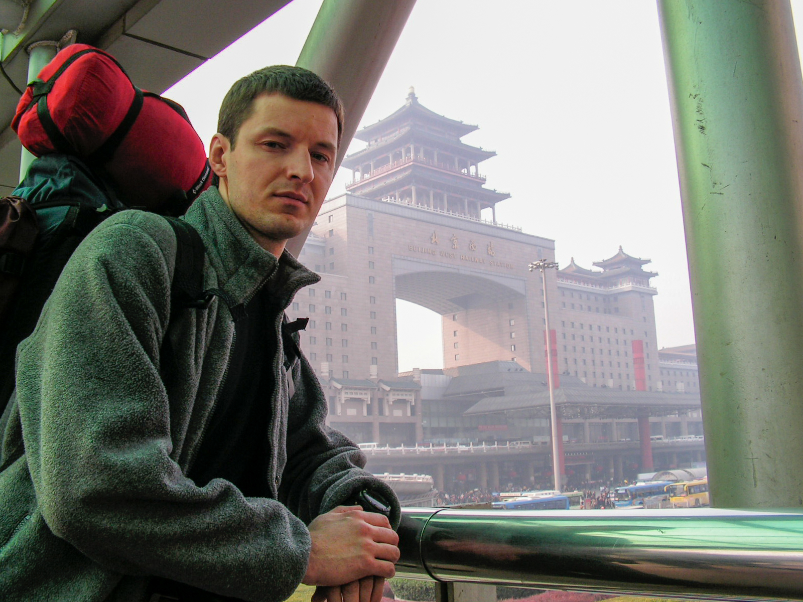 Kirill, a sinologist based in Beijing, in a grey fleece jacket with a large backpack and red stuff sack, leaning on a railing in front of Beijing West Railway Station in 2004.