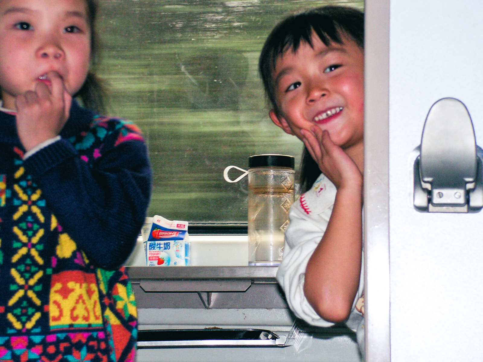 Two young Chinese girls at a train window table, one smiling shyly with her hand on her cheek and the other in a colourful patterned sweater, with a yoghurt carton and a tea flask between them.