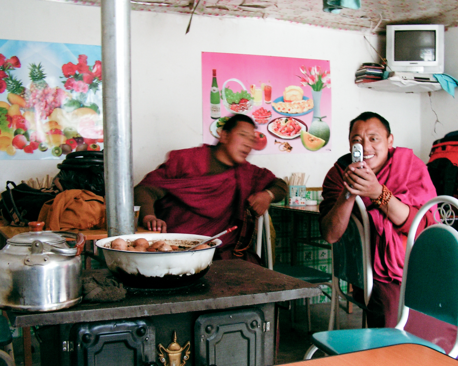 Two Tibetan monks in maroon robes sitting by a cast-iron stove in a small roadside eatery in 2004, one smiling and taking a photo with a flip phone, with a kettle and a bowl of eggs on the stove.
