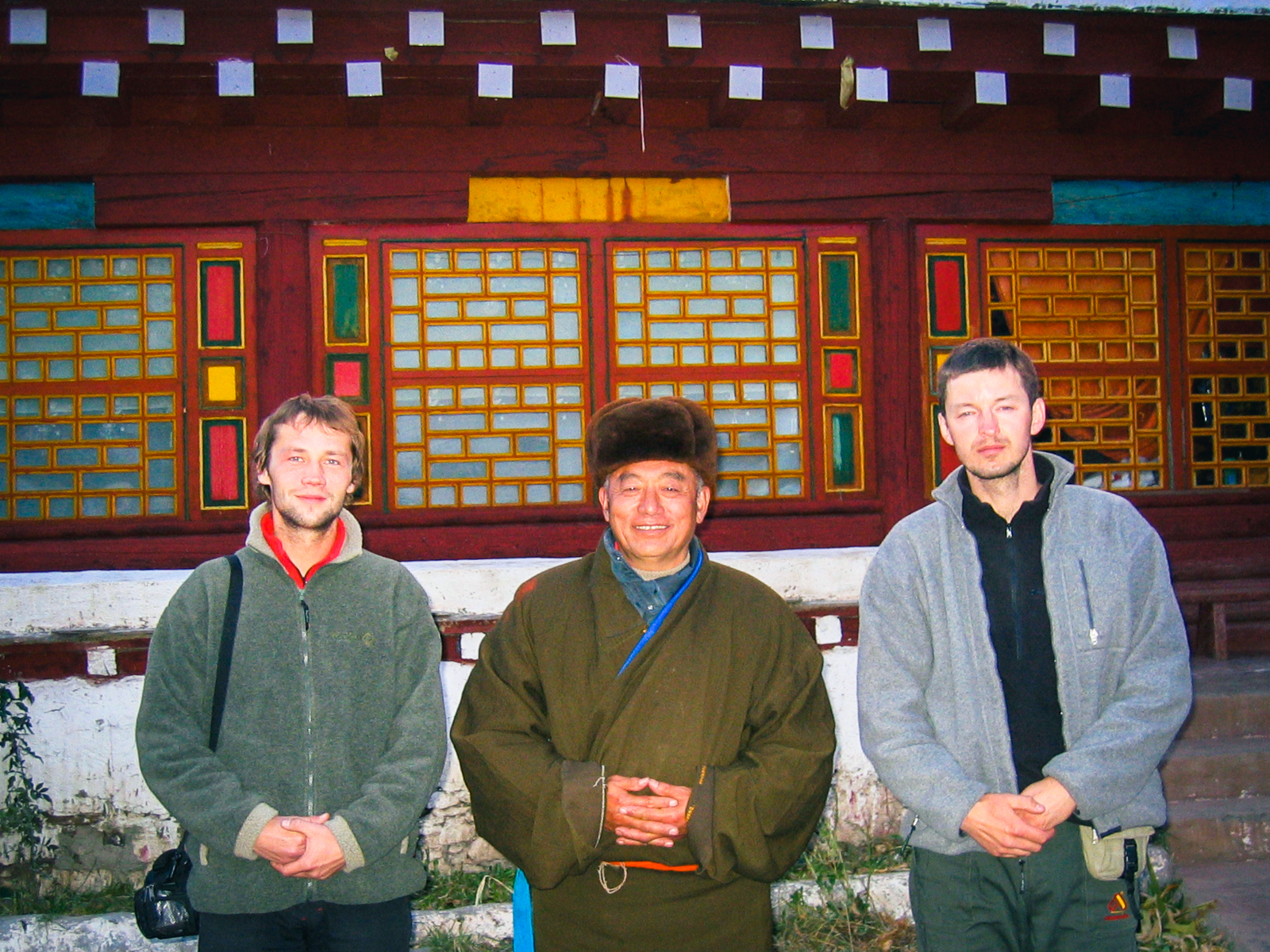 Oleg Gritskevich and his friend Kirill standing on either side of a smiling Tibetan man in a brown chuba robe and fur hat, in front of a monastery building with red, yellow and green painted lattice windows, in 2004.