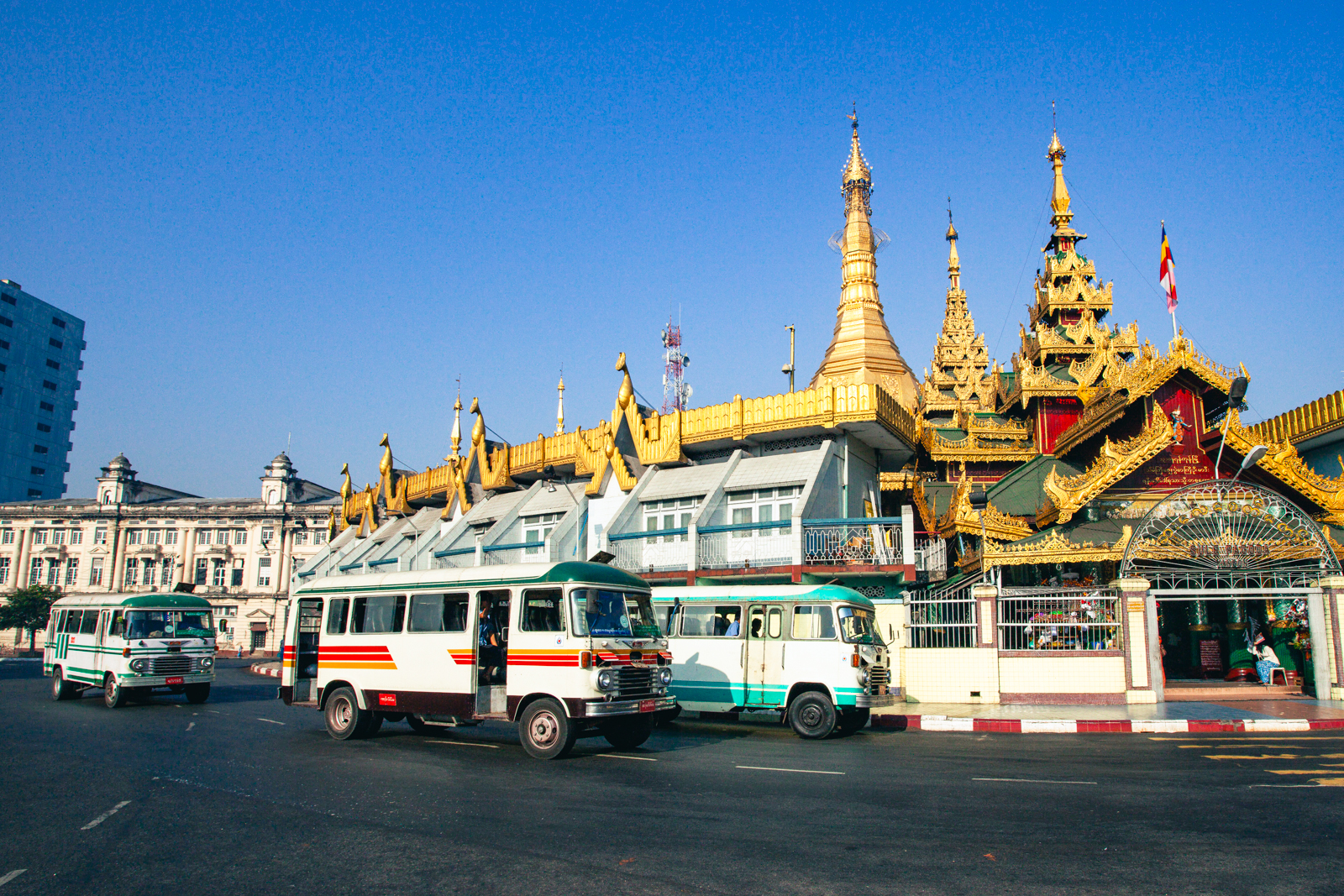 Vintage white buses with green and red stripes passing the golden Sule Pagoda on a roundabout in downtown Yangon, Myanmar, under a clear blue sky.