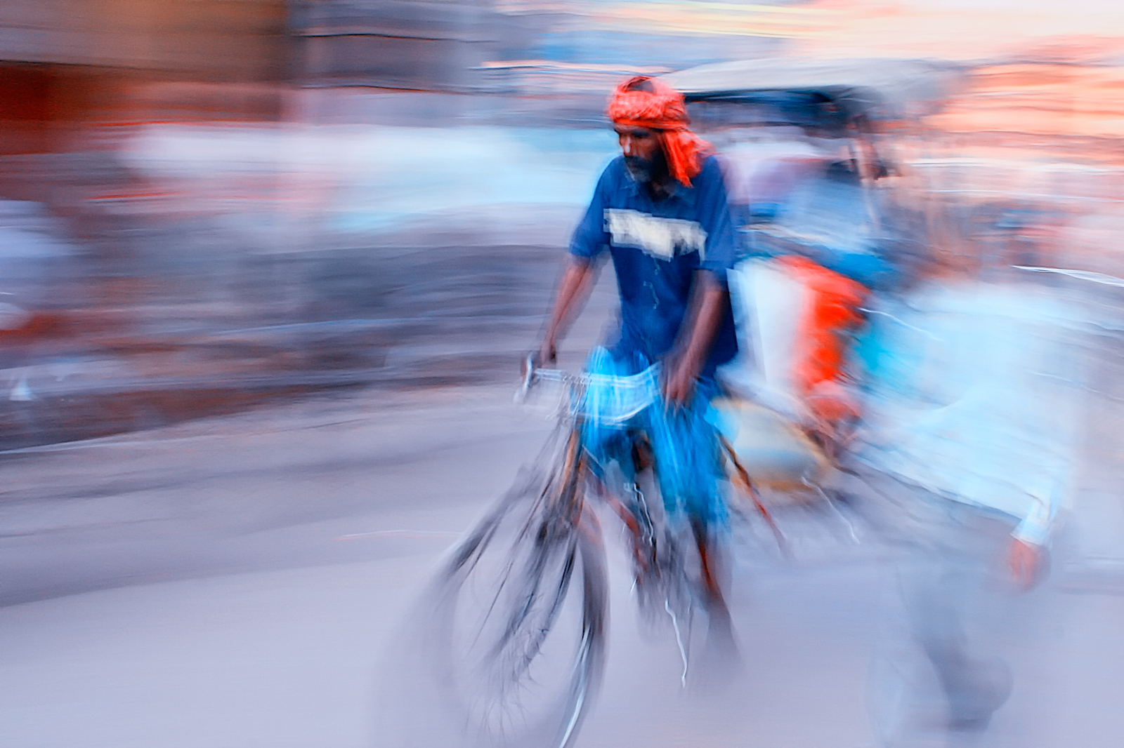 Cycle rickshaw driver in an orange headscarf and blue shirt riding through a street at dusk, captured with motion blur from a panning shot.